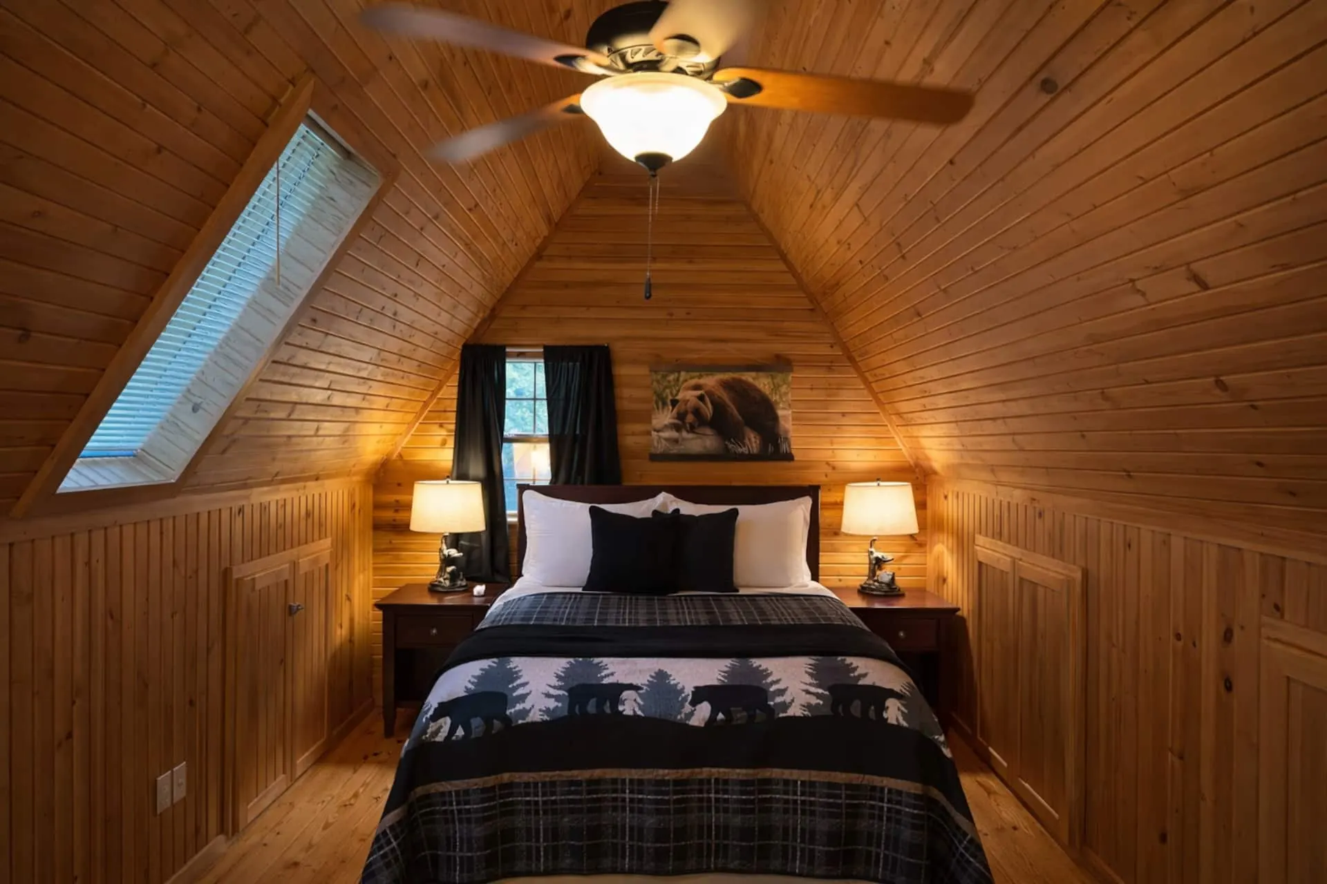 Upstairs bedroom with a queen bed under a vaulted pine ceiling, a skylight, bedside lamps and a ceiling fan