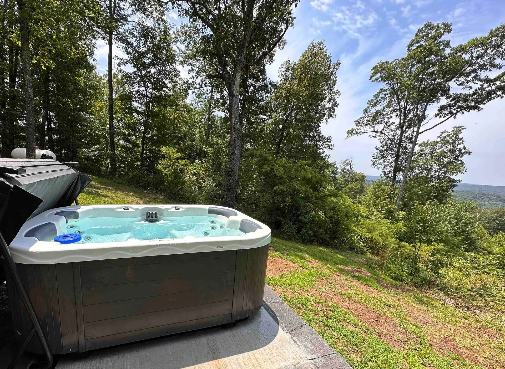 The private hot tub on its stone pad in daylight, with green Ozark mountain views and forest all around
