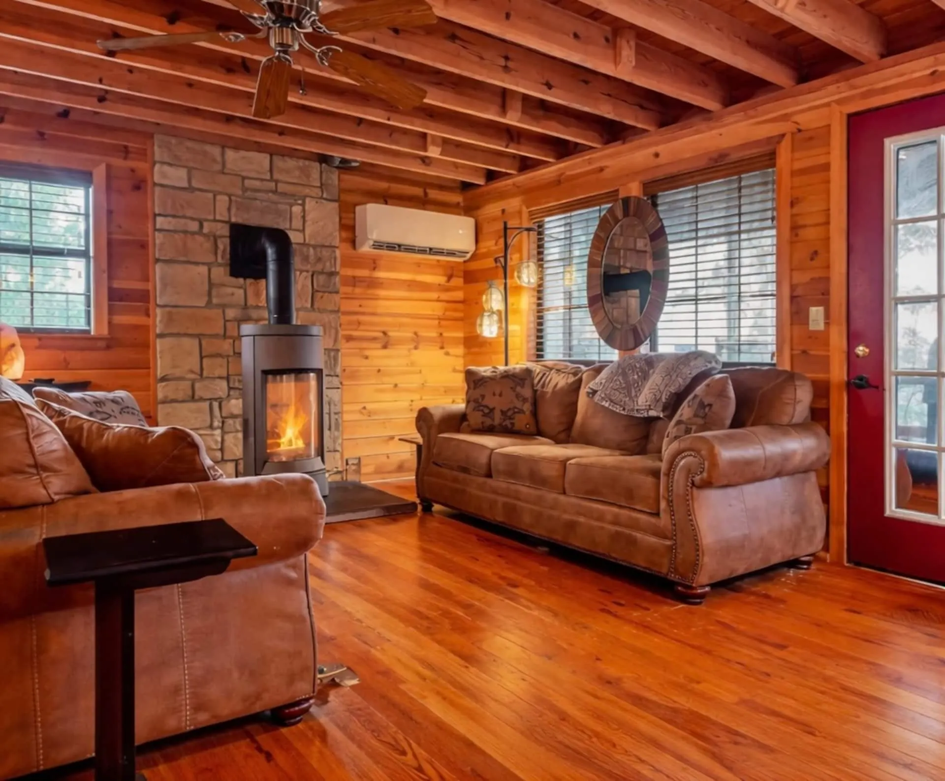 Leather sofa and loveseat in the log-walled living room beside the fireplace, with a red front door and hardwood floors
