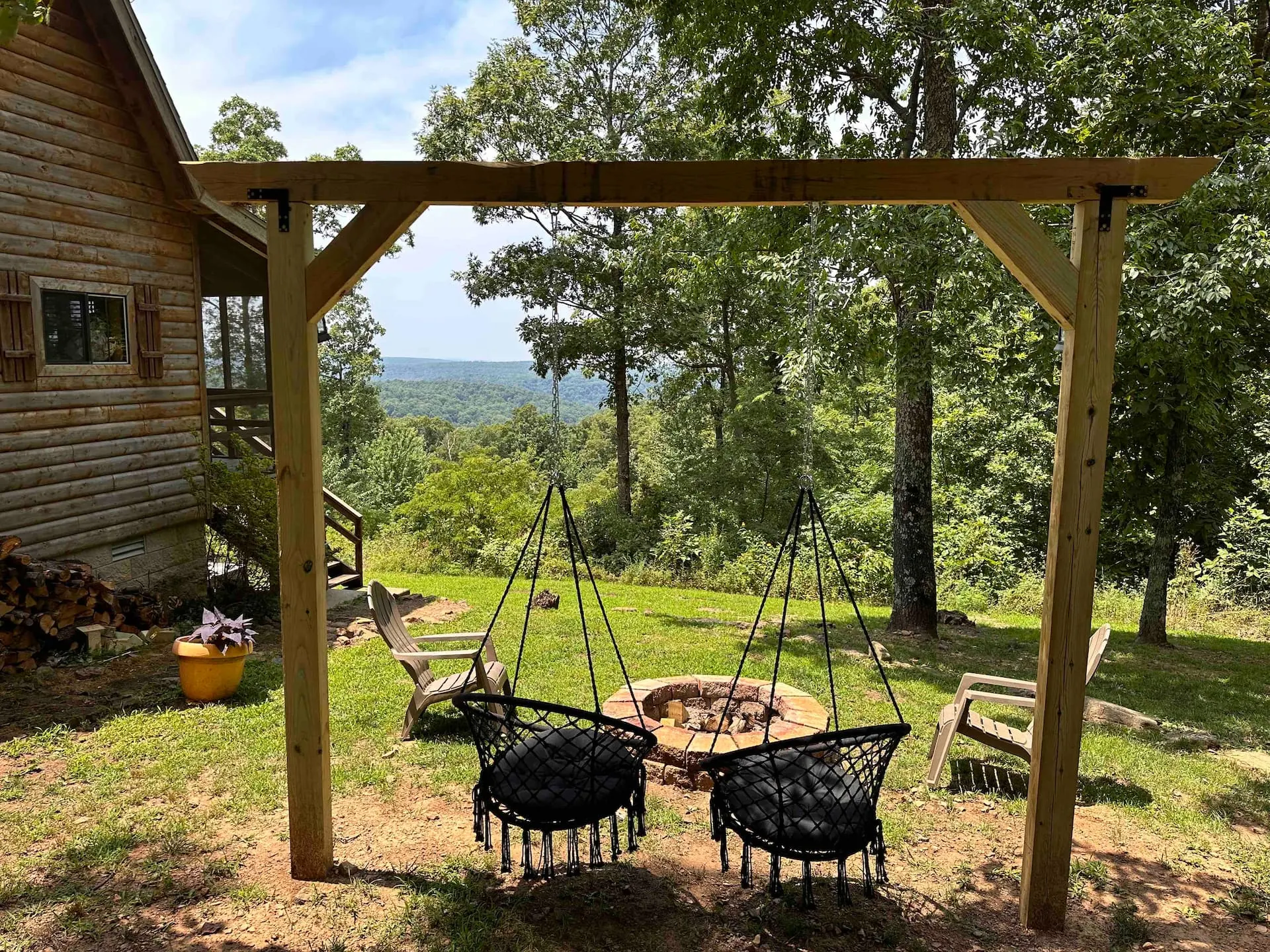 Two hanging swing chairs under a timber pergola beside the fire pit, with the cabin at left and the valley view ahead